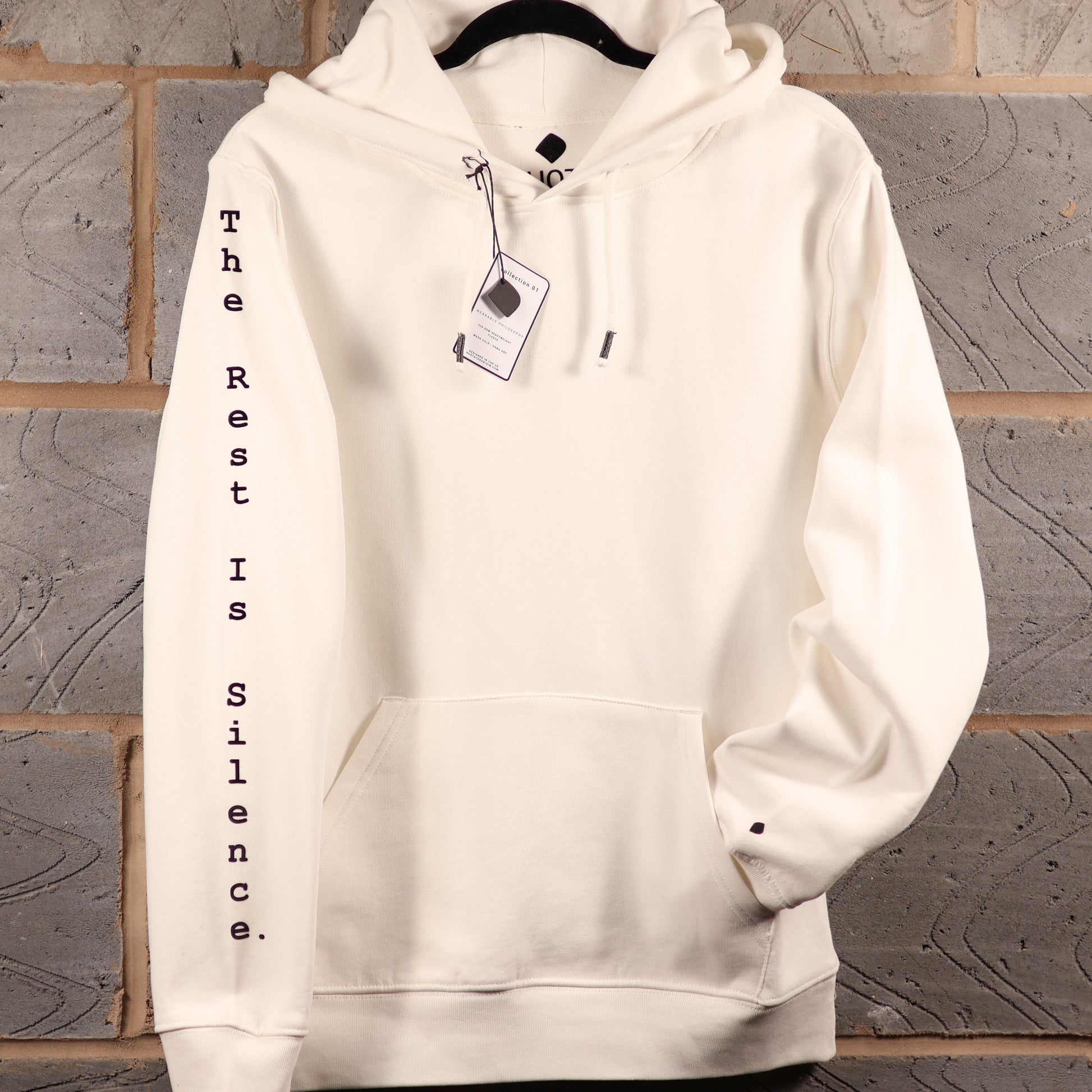 An off-white pullover hoodie hanging on a black hanger against a gray brick wall. The hoodie features 'The Rest Is Silence.