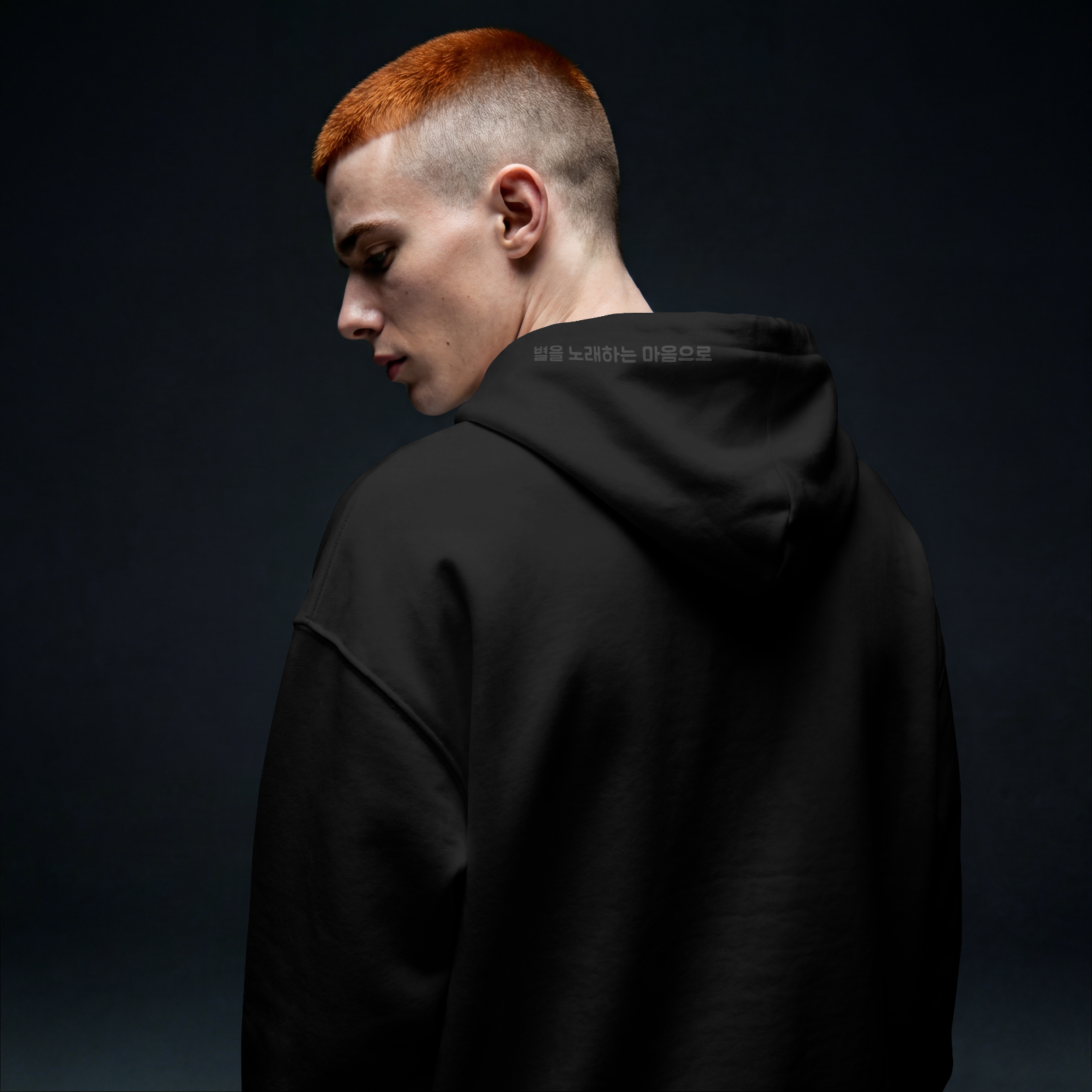 Model in dark studio lighting wearing a Nuqta black heavyweight hoodie, featuring subtle black-on-black Korean poetry typography on the hood. UK Brutalist streetwear.