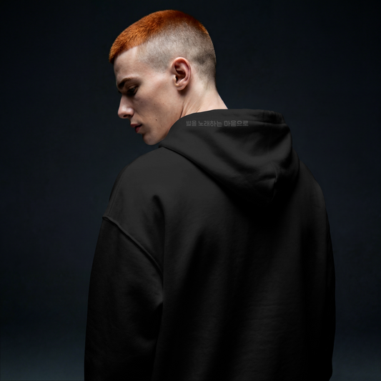 Model in dark studio lighting wearing a Nuqta black heavyweight hoodie, featuring subtle black-on-black Korean poetry typography on the hood. UK Brutalist streetwear.