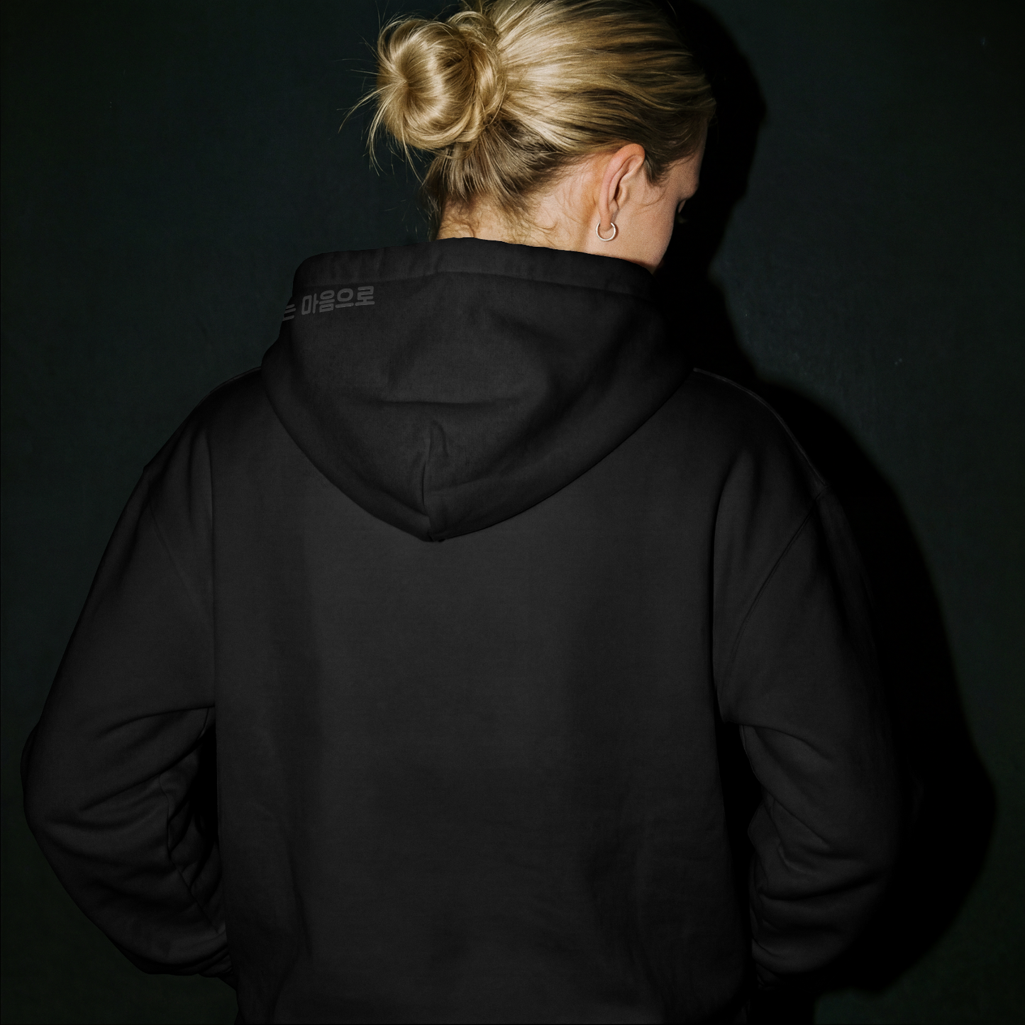 Model in dark studio lighting wearing a Nuqta black heavyweight hoodie, featuring subtle black-on-black Korean poetry typography on the hood. UK Brutalist streetwear.
