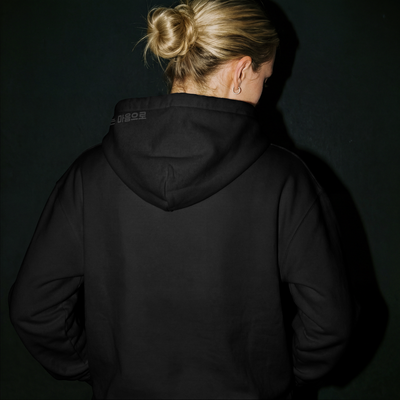 Model in dark studio lighting wearing a Nuqta black heavyweight hoodie, featuring subtle black-on-black Korean poetry typography on the hood. UK Brutalist streetwear.