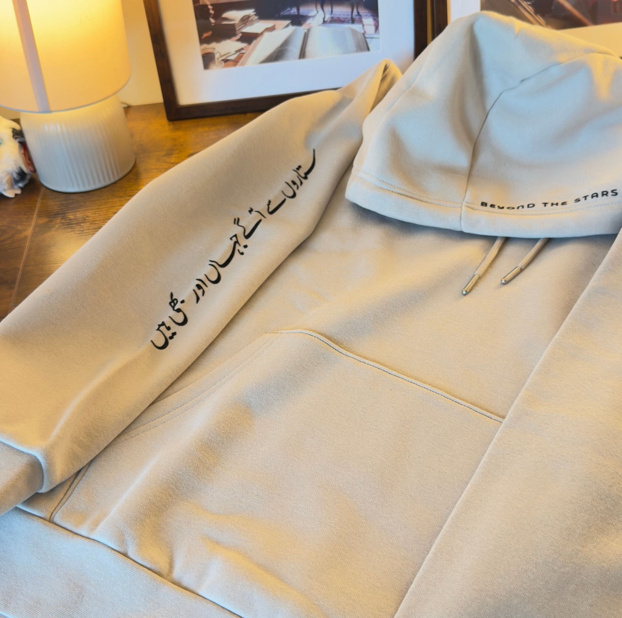 Beige parchment heavyweight fleece hoodie folded, showing black Urdu calligraphy on the sleeve.